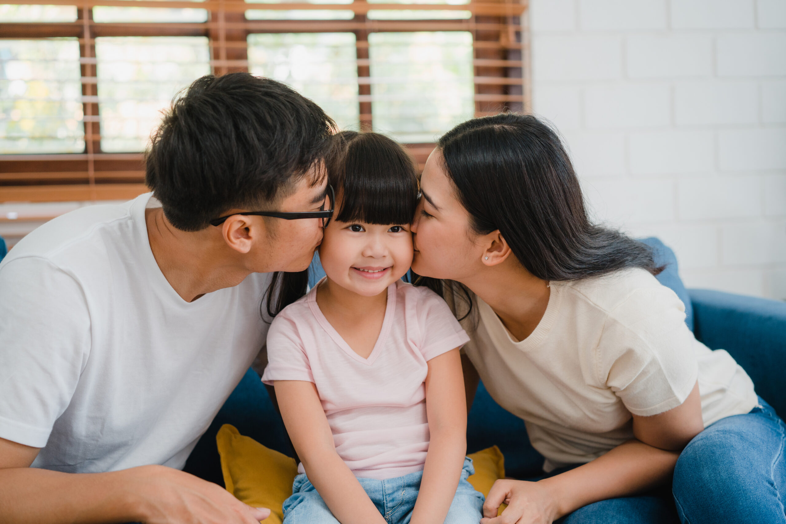A heartwarming scene of an Asian family on a couch, with parents kissing their daughter, showcasing love and togetherness.