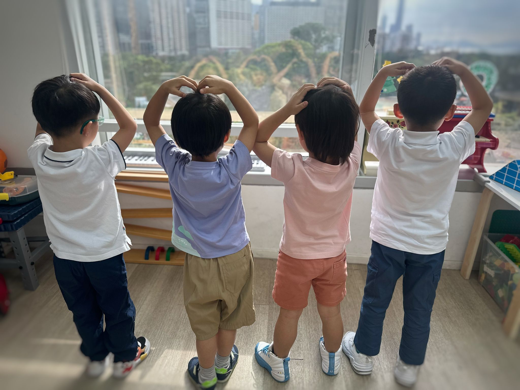 Four cheerful children pose with their arms forming a heart shape above their heads in front of a window, their faces reflecting in the glass.